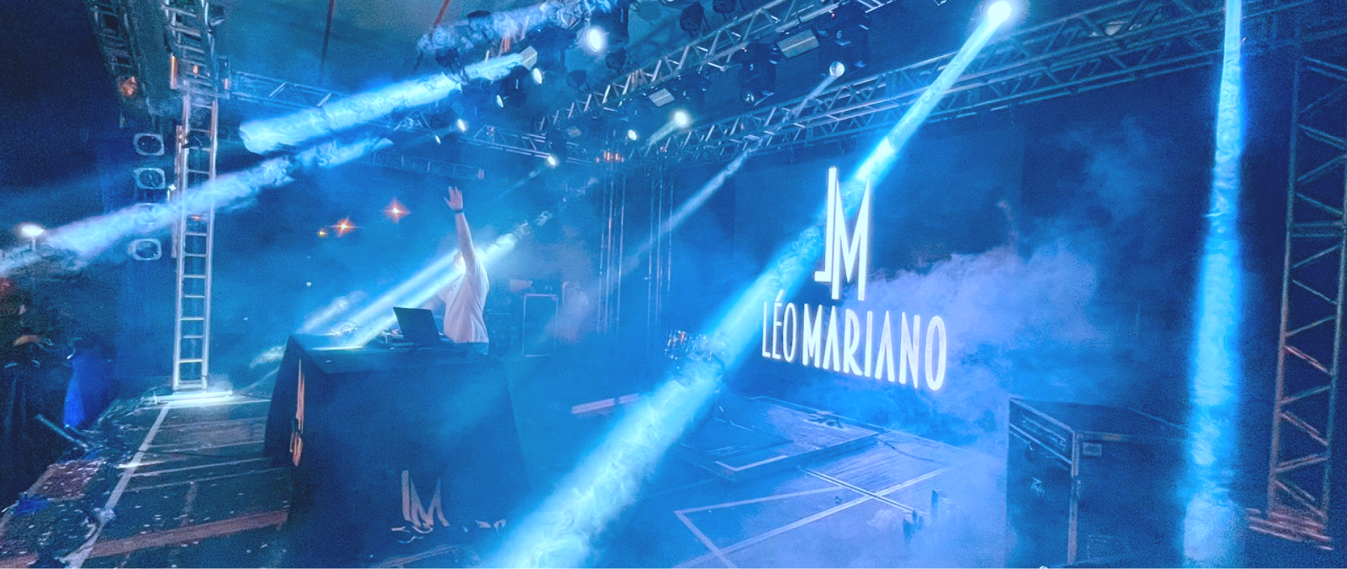 Imagem de DJ Leo Mariano com as mãos para o alto em um evento no palco.