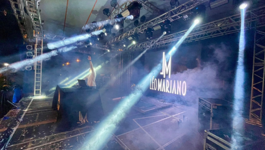 Imagem de DJ Leo mariano em um palco com a mão pro alto durante evento.