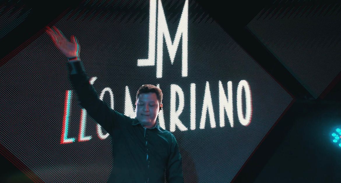 Imagem de DJ Leo Mariano com a mão para o alto em evento, e atras seu nome em uma tela.