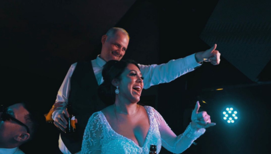 Imagem de um casal acenando para o DJ Leo Mariano, durante evento de casamento.